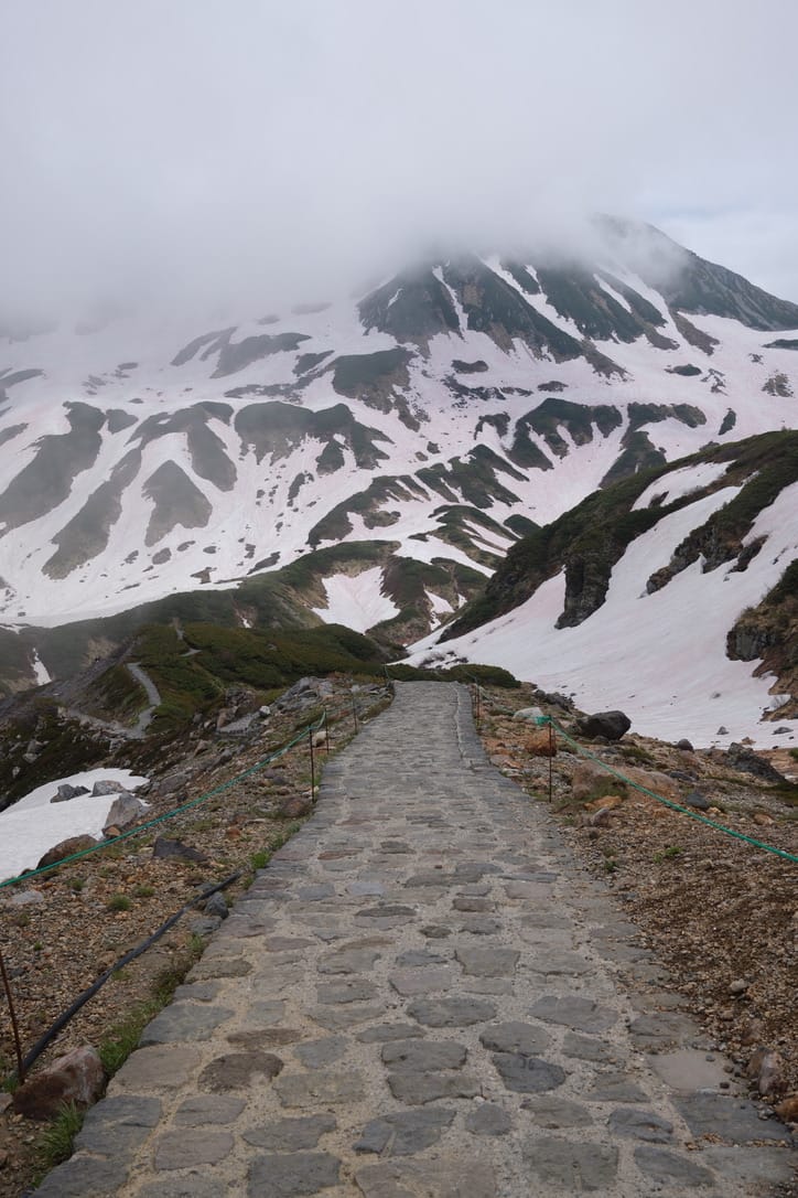 A cobbled path climbing through patchy snow toward a cloud-covered peak in the Japanese Alps