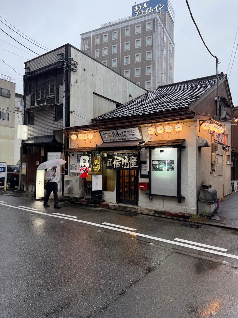 A small izakaya hung with paper lanterns on a wet street, a modern hotel rising behind it