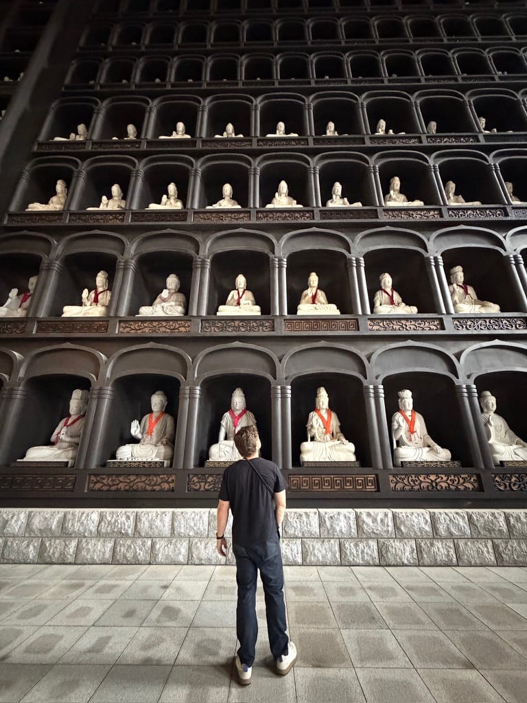 Andy Sommer standing in front of a towering wall of white seated Buddha statues at the Echizen Daibutsu