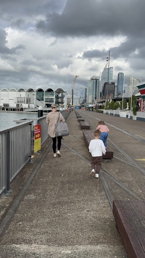 Children walking along a waterfront promenade with a city skyline behind them.