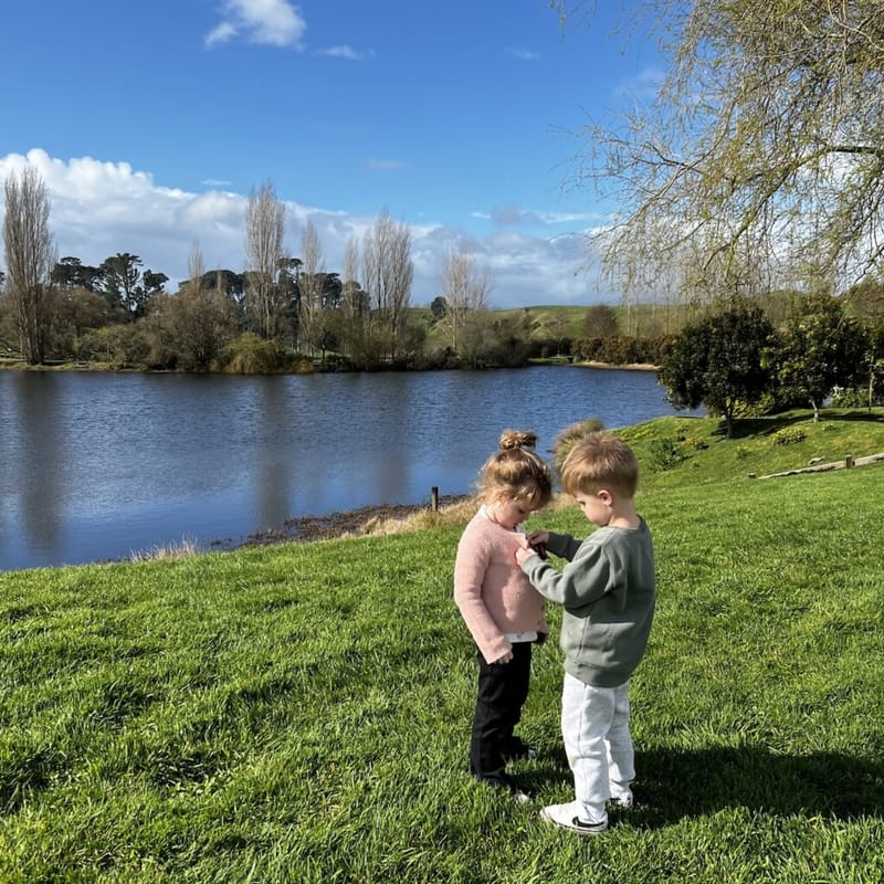 Two children standing by a lake among rolling green hills.